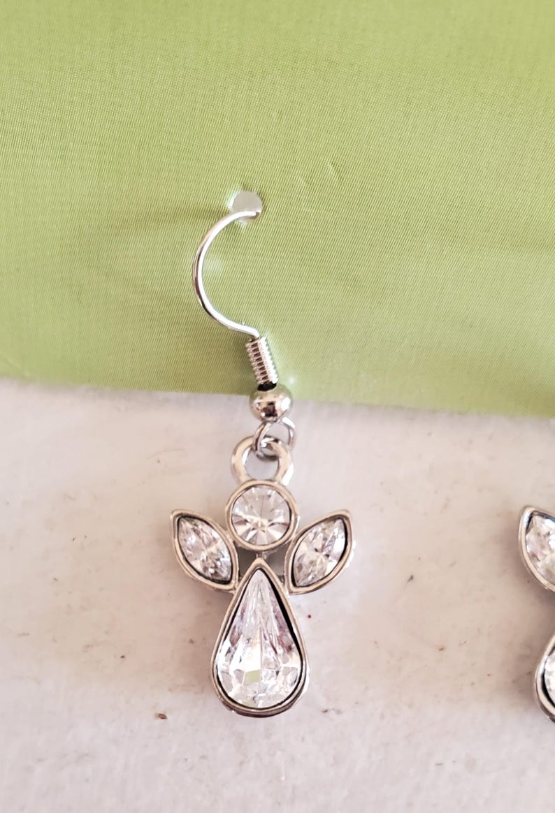 Puede incluir: Pendientes de &aacute;ngel plateados con cristales transparentes y brillantes. Cada pendiente presenta un cristal en forma de l&aacute;grima en el centro, rodeado de cristales m&aacute;s peque&ntilde;os con forma de marquesa y un cristal redondo en la parte superior. Cierre de gancho.