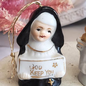 Puede incluir: Figura de cer&aacute;mica de una monja con h&aacute;bito negro y toca blanca. Sostiene un letrero con el texto dorado "GOD KEEP YOU" y una peque&ntilde;a cruz dorada. Tiene un cord&oacute;n dorado para colgar.