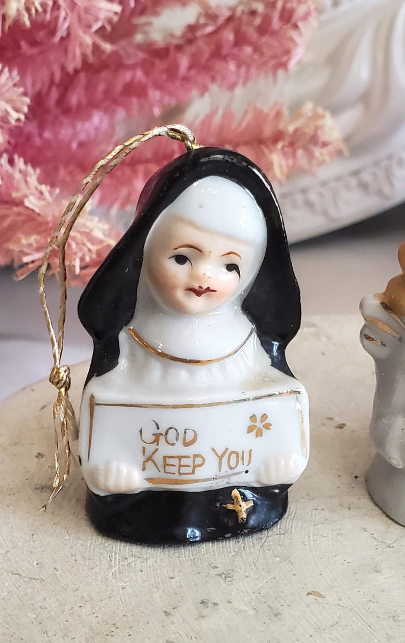 Puede incluir: Figura de cer&aacute;mica de una monja con h&aacute;bito negro y toca blanca. Sostiene un letrero con el texto dorado "GOD KEEP YOU" y una peque&ntilde;a cruz dorada. Tiene un cord&oacute;n dorado para colgar.