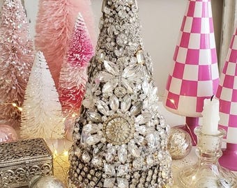 May include: A sparkling Christmas tree made of crystals and rhinestones, surrounded by pink bottle brush trees and decorative ornaments. The tree is adorned with large crystal embellishments and sits on a white surface. A vintage-style holiday display.