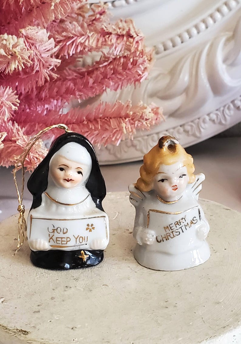 Puede incluir: Dos adornos navide&ntilde;os de cer&aacute;mica. Uno es una monja que sostiene un letrero que dice "God Keep You". El otro es un &aacute;ngel que sostiene un letrero que dice "Merry Christmas". Ambos son blancos con detalles dorados.