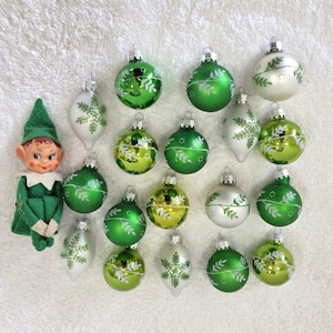 May include: A green elf doll with a pointed hat and white collar positioned next to a collection of fifteen Christmas ornaments. The ornaments include various round and teardrop-shaped glass baubles in shades of green, lime, and white.