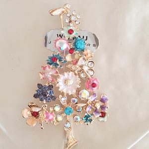 May include: A gold-tone brooch shaped like a Christmas tree, decorated with colorful enamel flowers, rhinestones, and a dragonfly at the top. The brooch has a tag that reads "Willau Jewelers".
