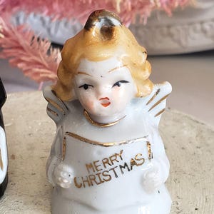 Puede incluir: Peque&ntilde;a figura de &aacute;ngel de cer&aacute;mica blanca con adornos dorados y cabello rubio. El &aacute;ngel sostiene una pancarta que dice "MERRY CHRISTMAS" en letras doradas. La figura tiene peque&ntilde;as alas y es una decoraci&oacute;n navide&ntilde;a.
