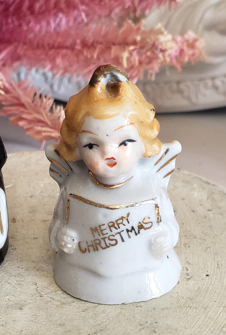 Puede incluir: Peque&ntilde;a figura de &aacute;ngel de cer&aacute;mica blanca con adornos dorados y cabello rubio. El &aacute;ngel sostiene una pancarta que dice "MERRY CHRISTMAS" en letras doradas. La figura tiene peque&ntilde;as alas y es una decoraci&oacute;n navide&ntilde;a.