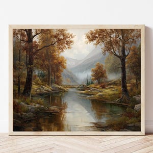 Op de afbeelding: Een ingelijst landschapsschilderij met een serene rivierscène. Het kunstwerk toont bomen met herfstkleuren in bruin en goud, die weerspiegeld worden in het kalme water. Bergen zijn zichtbaar op de achtergrond, met een zachte, nevelige sfeer.