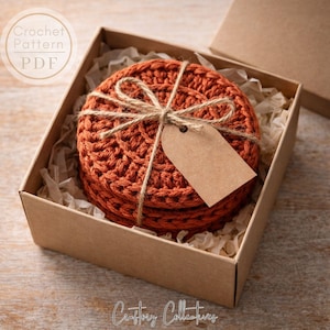 Puede incluir: Un conjunto de posavasos de ganchillo naranja, atados con hilo y una etiqueta en blanco, presentados en una caja de cartón con papel. La caja tiene el texto "Crochet Pattern PDF" y "Craftory Collectives".