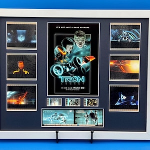 以下が含まれることがあります： 映画「トロン：レガシー」のフレーム入りポスターとフィルムセル。ポスターにはキャラクターとライトサイクルが描かれています。フレームには映画のシーンが映ったフィルムセルが含まれています。テキストは「IT'S NOT JUST A GAME ANYMORE」と「TRON LEGACY IN 3D AND IMAX 3D」と書かれています。