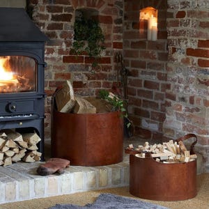 Peut inclure: Deux paniers à bûches cylindriques en cuir marron dotés de poignées robustes, remplis de bois de chauffage fendu. Les paniers sont placés sur un âtre en pierre près d'un poêle à bois en métal noir allumé, devant un mur en briques rouges.