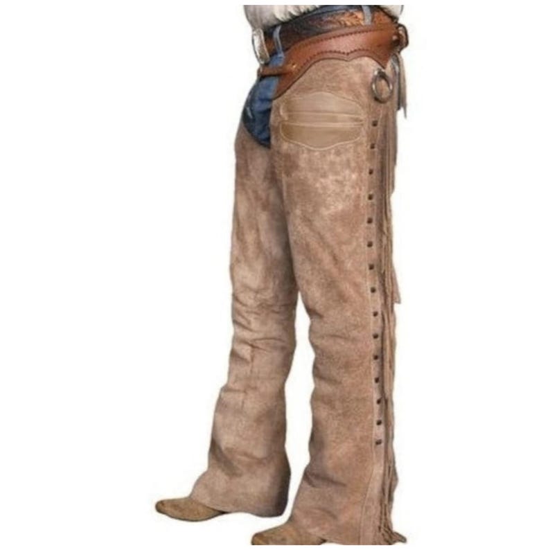 May include: Tan suede leather chaps featuring a dark brown leather waistband with a buckle closure, a smooth leather pocket detail on the upper thigh, and long fringe trim running down the outer side of each leg over blue denim jeans.