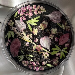 Puede incluir: Una bandeja redonda de resina transparente con base negra, que muestra una disposición de flores y follaje secos. Los elementos botánicos incluyen pétalos rosas y blancos, hojas verdes y delicados tallos. La bandeja se encuentra sobre un fondo blanco y sedoso.