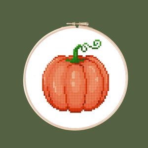 Puede incluir: Diseño de punto de cruz de una calabaza en un bastidor de bordado. La calabaza es naranja con un tallo y una vid verdes. El fondo es de color verde oliva liso. El texto "Kitty Kross Stitch" está en un círculo de huellas de patas.