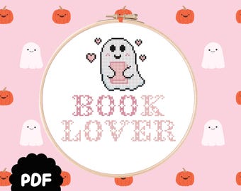 May include: A cross-stitch pattern in a wooden embroidery hoop featuring a white ghost holding a pink book, surrounded by four small pink hearts. Below the ghost, the text reads BOOK LOVER in pink pixelated letters. A black PDF icon is visible.