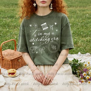 May include: Olive green t-shirt with the text "In my stitching era" and white illustrations of sewing tools and floral designs. The shirt is a relaxed fit, perfect for crafters and those who enjoy embroidery or cross-stitch.