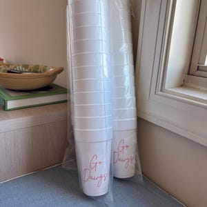 Puede incluir: Dos pilas de vasos desechables blancos, cada uno envuelto en plástico transparente. Las tazas tienen una escritura roja que dice "Go Dawgs". Las tazas están apiladas verticalmente, listas para usar.