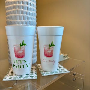 Könnte beinhalten: Zwei weiße Schaumstoffbecher mit einem Aquarell-Cocktail-Design und dem Text "LET'S PARTY!" und "Let's Party!". Die Becher stehen auf gemusterten Untersetzern, passend für eine Party.