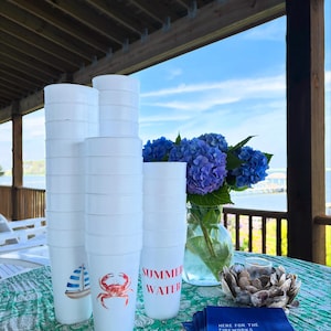 May include: A stack of white plastic cups with nautical-themed designs, including a sailboat and a crab, and the words "SUMMER WATER." A vase of blue hydrangeas and a stack of blue napkins with the text "HERE FOR THE FIREWORKS" are also visible.