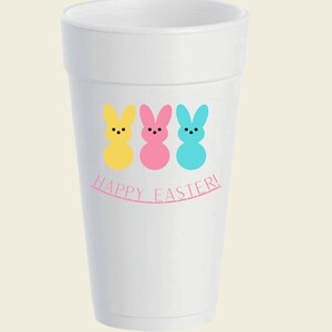 May include: A white foam cup featuring three colorful bunny designs in yellow, pink, and blue. The words "Happy Easter!" are printed in pink below the bunnies, making it a festive item for Easter celebrations.