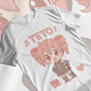以下が含まれることがあります： 白いTシャツ。ピンクの髪と黒と赤の衣装を着たキャラクターの漫画イラスト。キャラクターの上には「TETO！」の文字と音符があります。ピンクのハートと数字「040」も表示されています。