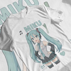 以下が含まれることがあります： 白いTシャツ。長い水色のツインテールで、制服を着たキャラクターの漫画イラストがプリントされています。「MIKU」の文字、音符、ネギが描かれています。ハートも表示されています。