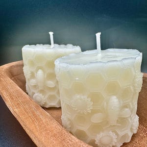 Puede incluir: Dos velas cilíndricas de color blanquecino con un diseño de panal y abeja, en un cuenco de madera. Las velas tienen una superficie texturizada y una mecha en la parte superior. El fondo es de color verde azulado oscuro.