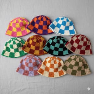 Op de afbeelding: Een collectie van tien gehaakte bucket hats gerangschikt in drie rijen. Elke hoed heeft een opvallend tweekleurig ruitpatroon in diverse kleurcombinaties, waaronder roze en geel, paars en oranje, blauw en wit, groen en wit, bruin en geel, zwart en groenblauw, bordeaux en wit, lavendel en paars, oranje en crème, en olijf en beige.