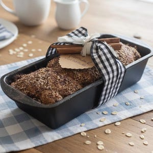 May include: A loaf of bread in a black rectangular baking pan, topped with seeds and spices. The bread is wrapped with a black and white checkered ribbon and a cinnamon stick. A tag reads "Oatmeal Cinnamon".