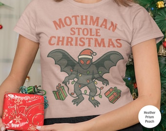 Op de afbeelding: Perzikkleurig T-shirt met de tekst "Mothman Stole Christmas". De afbeelding toont een Mothman-figuur met een kerstmuts en lichtjes, met ingepakte cadeaus. Een grappig en feestelijk ontwerp.