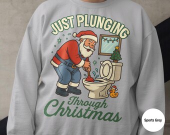 Pode incluir: Camisola cinza desportiva com um gráfico de Natal humorístico. O design apresenta o Pai Natal a usar um desentupidor numa sanita, com o texto "Just Plunging Through Christmas" em verde. Inclui também uma pequena árvore de Natal e um patinho de borracha.