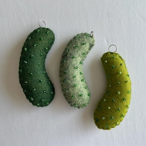 Puede incluir: Tres adornos de fieltro con forma de pepinillo en varios tonos de verde, incluyendo verde bosque oscuro, salvia y verde lima brillante. Cada pieza de fieltro curvada y texturizada est&aacute; decorada con peque&ntilde;as cuentas verdes y una argolla plateada.