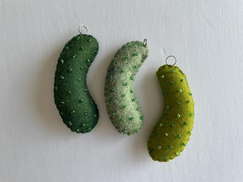 Puede incluir: Tres adornos de fieltro con forma de pepinillo en varios tonos de verde, incluyendo verde bosque oscuro, salvia y verde lima brillante. Cada pieza de fieltro curvada y texturizada est&aacute; decorada con peque&ntilde;as cuentas verdes y una argolla plateada.