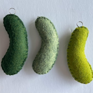 Puede incluir: Tres adornos de fieltro curvados en varios tonos de verde, incluyendo verde bosque oscuro, salvia apagado y lima brillante. Cada pieza presenta puntadas de fest&oacute;n visibles a lo largo de los bordes y una peque&ntilde;a anilla de metal plateado arriba.
