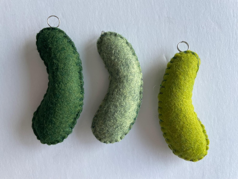 Puede incluir: Tres adornos de fieltro curvados en varios tonos de verde, incluyendo verde bosque oscuro, salvia apagado y lima brillante. Cada pieza presenta puntadas de fest&oacute;n visibles a lo largo de los bordes y una peque&ntilde;a anilla de metal plateado arriba.