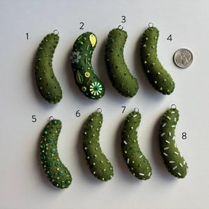 Puede incluir: Ocho adornos de pepinillos de fieltro verde numerados del uno al ocho, cada uno con un peque&ntilde;o lazo met&aacute;lico para colgar. Los pepinillos presentan varios adornos de cuentas y lentejuelas. Se incluye una moneda para la escala.