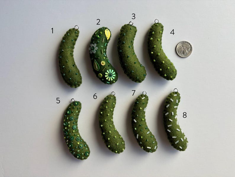 Puede incluir: Ocho adornos de pepinillos de fieltro verde numerados del uno al ocho, cada uno con un peque&ntilde;o lazo met&aacute;lico para colgar. Los pepinillos presentan varios adornos de cuentas y lentejuelas. Se incluye una moneda para la escala.