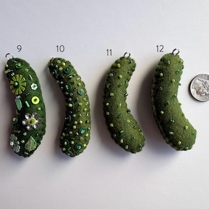 Puede incluir: Cuatro adornos de pepinillo de fieltro verde oliva etiquetados 9, 10, 11 y 12, cada uno con adornos &uacute;nicos de cuentas y botones verdes. Un anillo de metal plateado est&aacute; unido a la parte superior de cada uno. Una moneda de diez centavos sirve de escala.