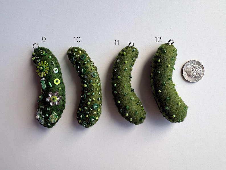 Puede incluir: Cuatro adornos de pepinillo de fieltro verde oliva etiquetados 9, 10, 11 y 12, cada uno con adornos &uacute;nicos de cuentas y botones verdes. Un anillo de metal plateado est&aacute; unido a la parte superior de cada uno. Una moneda de diez centavos sirve de escala.