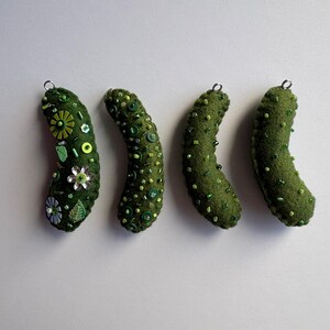 Puede incluir: Cuatro adornos de fieltro verde con forma de pepinillo, cada uno con una peque&ntilde;a anilla met&aacute;lica en la parte superior. Los adornos est&aacute;n decorados con diversas cuentas verdes y transparentes, lentejuelas y peque&ntilde;os botones florales.