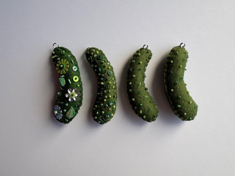 Puede incluir: Cuatro adornos de fieltro verde con forma de pepinillo, cada uno con una peque&ntilde;a anilla met&aacute;lica en la parte superior. Los adornos est&aacute;n decorados con diversas cuentas verdes y transparentes, lentejuelas y peque&ntilde;os botones florales.