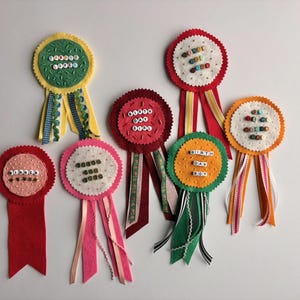 May include: A collection of handmade felt rosettes in various colors, including red, yellow, and pink. Each rosette features a circular design with text and decorative ribbons. The text on the rosettes includes phrases like "Good Job" and "Birth Day Boy."