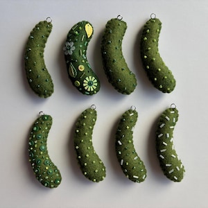 Puede incluir: Ocho adornos de fieltro en forma de pepinillo de color verde oliva dispuestos en dos filas. Cada pepinillo presenta adornos &uacute;nicos que incluyen varias cuentas verdes, lentejuelas y cuentas de semillas blancas. Cada adorno tiene un peque&ntilde;o aro de metal plateado.