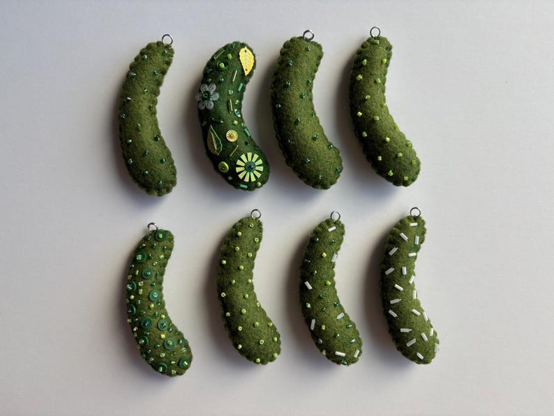 Puede incluir: Ocho adornos de fieltro en forma de pepinillo de color verde oliva dispuestos en dos filas. Cada pepinillo presenta adornos &uacute;nicos que incluyen varias cuentas verdes, lentejuelas y cuentas de semillas blancas. Cada adorno tiene un peque&ntilde;o aro de metal plateado.