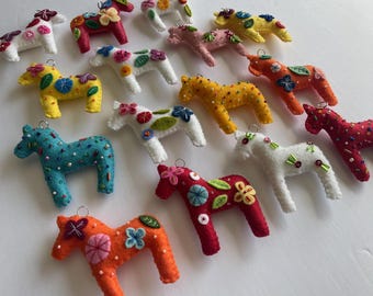May include: A collection of felt horse ornaments in various colors including red, orange, yellow, white, and blue. Each horse features intricate hand-stitched floral embroidery, small beads, and a metal loop at the top for hanging purposes.
