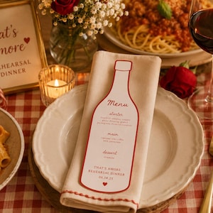 Könnte beinhalten: Eine weiße Menükarte in Form einer Weinflasche mit rotem Text und rotem Rand, die auf einer gefalteten beigen Serviette auf einem weißen Teller liegt. Das Menü enthält den Text Menu, starter, main, dessert und That's Amore Rehearsal Dinner 06.14.25.