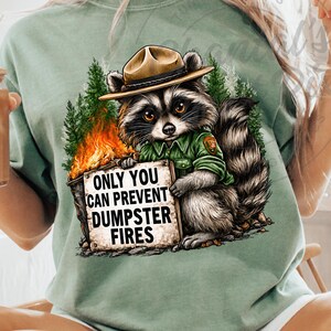 Puede incluir: Camiseta verde salvia con un mapache con uniforme de guardabosques, que lleva un sombrero y sostiene un letrero que dice "ONLY YOU CAN PREVENT DUMPSTER FIRES". El mapache está sobre un fondo de llamas y árboles.