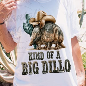 May include: White t-shirt featuring a detailed illustration of an armadillo wearing a cowboy hat, with a cactus in the background. The text "KIND OF A BIG DILLO" is printed below the armadillo in a Western-style font.