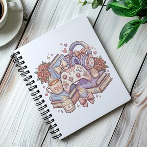 May include: Spiral-bound notebook featuring a pastel illustration of a gaming controller, headphones, books, coffee mug, candles, and roses. The design incorporates hearts and bows, creating a whimsical and cozy aesthetic. The notebook is on a white wooden surface.