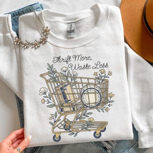 May include: White sweatshirt with a graphic of a shopping cart filled with books and a plate, surrounded by floral elements. The text "Thrift More, Waste Less" is above the cart. A floral necklace and jeans are in the background.