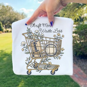 May include: White sweatshirt featuring a gold shopping cart graphic filled with books and plates, adorned with floral accents. The text "Thrift More, Waste Less" is written above the cart. The sweatshirt is held up by a person.