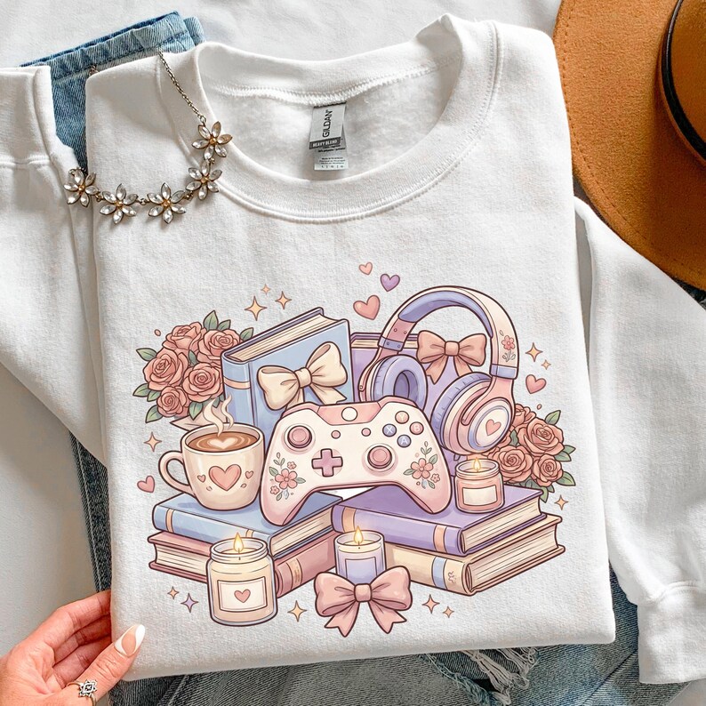 May include: White sweatshirt featuring a pastel illustration of a video game controller, books, headphones, coffee cup, candles, and roses. The design includes bows, hearts, and floral accents, creating a cozy, aesthetic theme.